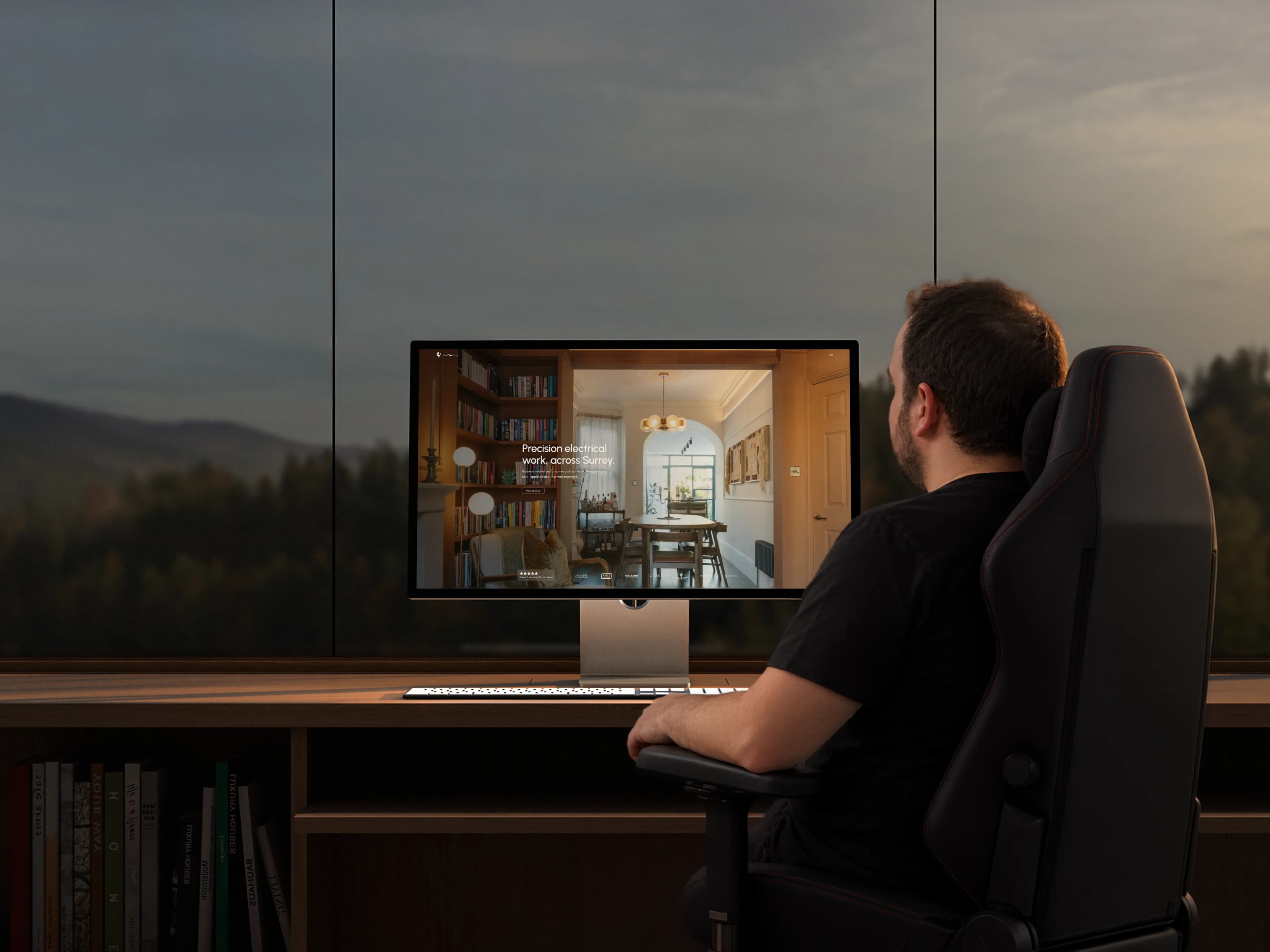 Michael at his desk at dusk, the JustElectrix website on a large monitor, hills beyond the window